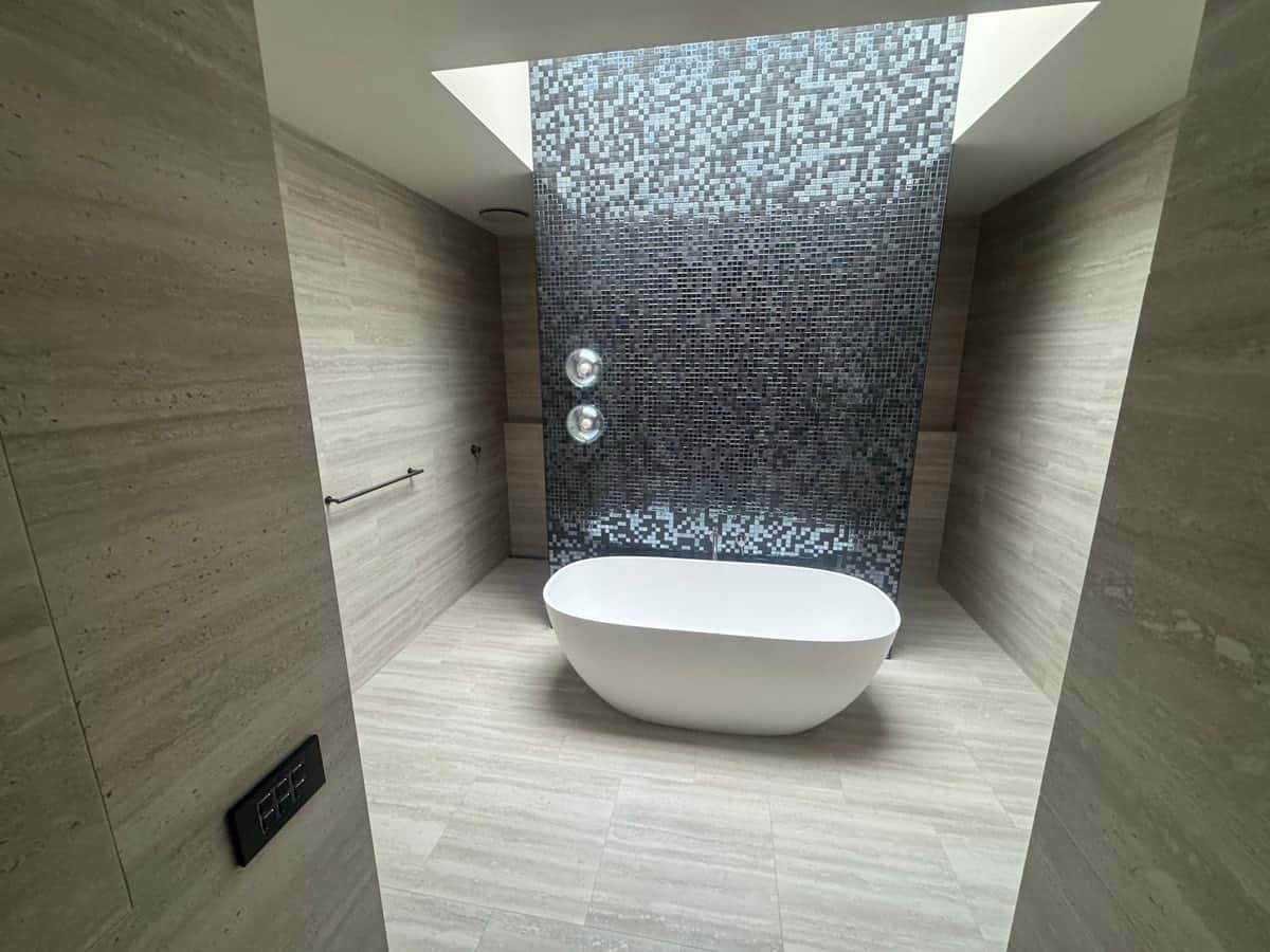 Freestanding bath set in front of a dark mosaic feature wall under a skylight.